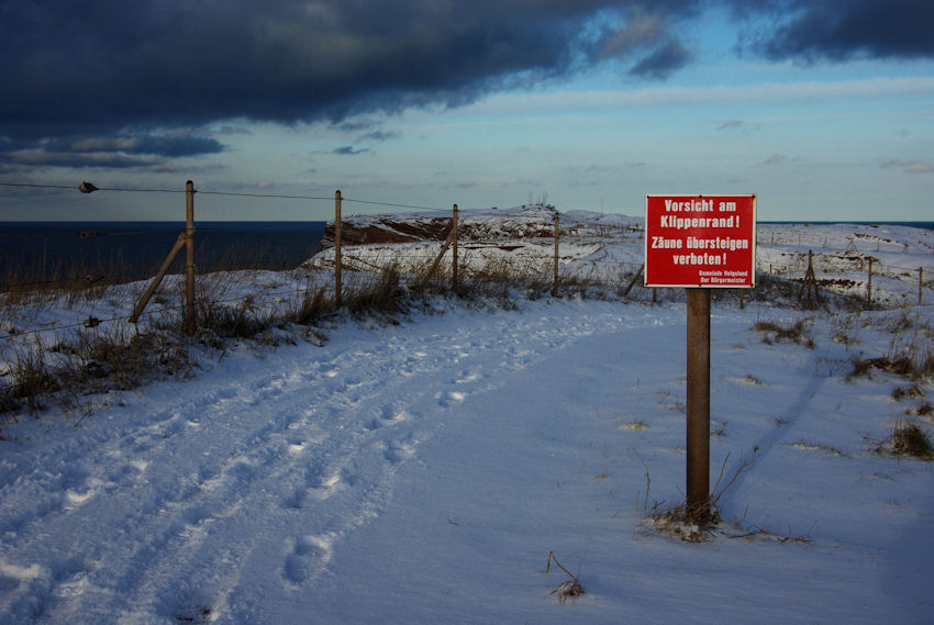 Am verschneiten Klippenrandweg Helgolands ist das leuchtend rote Schild mit weißer Schrift:"Vorsicht am Klippenrandweg! Zäune übersteigen verboten. Gemeinde Helgoland Der Bürgermeister" aufgestellt.