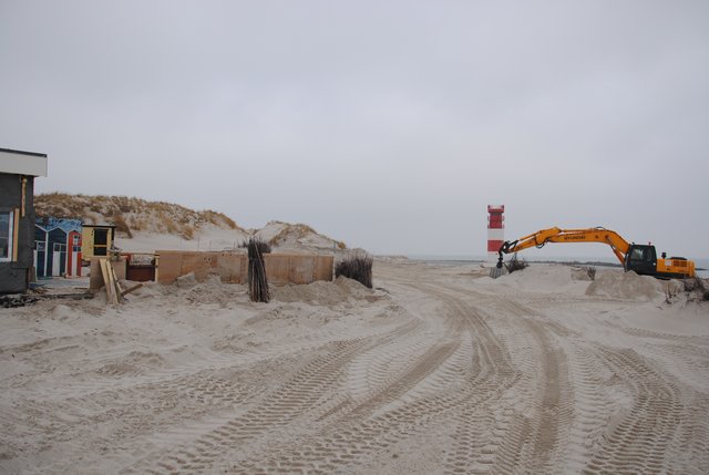Blick am Dünenrestaurant vorbei zum rot-weiß geringeltem Leuchtturm Oberfeuer Düne mit einem Raupenbagger am Strand. 