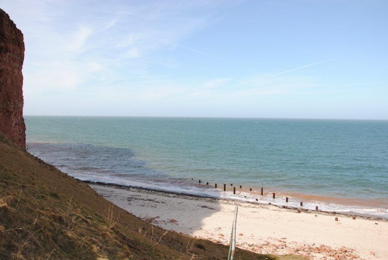 Von halber Höhe des Helgoländer Jägerstiegs Richtung Norden fotografiert, mit dem Schatten des Felsens, der sich auf dem weißen Strand und dem blaugrünen Meer zeigt.