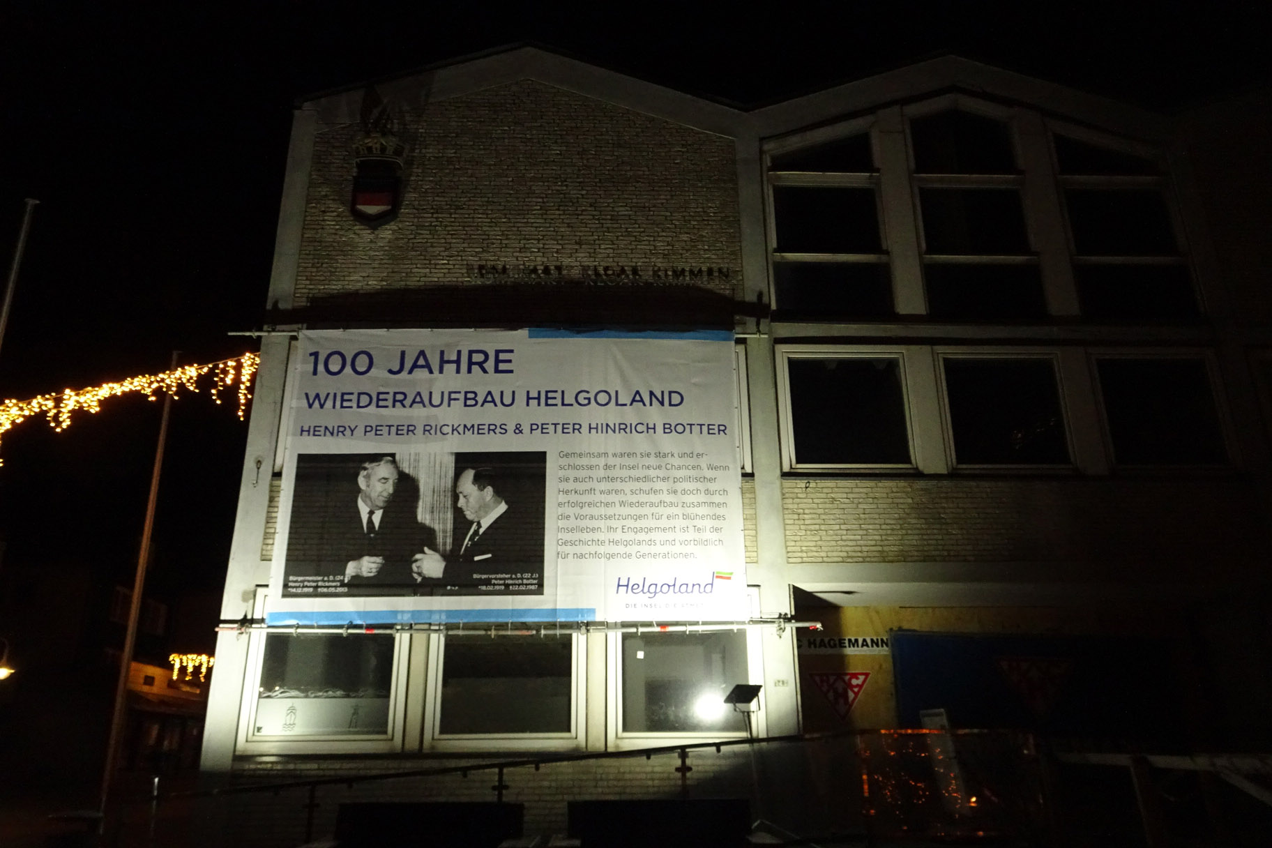 Die Eingangsseite des Helgoländer Rathauses bei Dunkelheit mit einem Riesen-Plakat zum 100. Geburtstag von H.P. Rickmers und P.H. Botter.