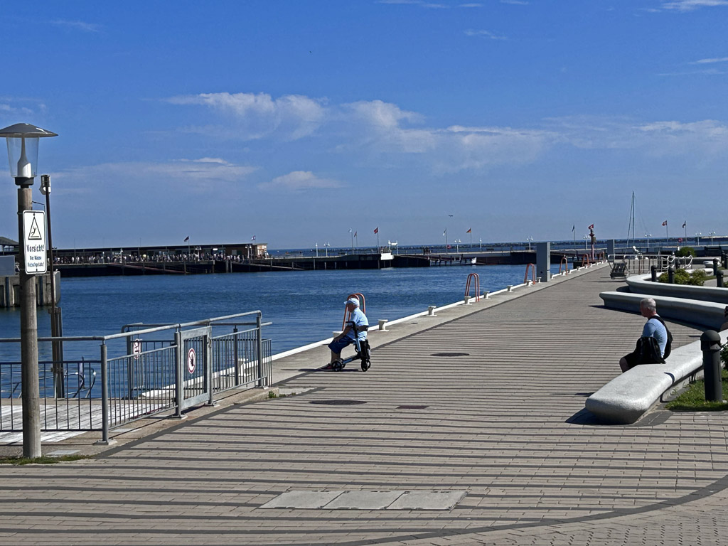 Blick von der Hafenstra&szlig;e auf Helgoland durch die Scheibenhafenausfahrt hin&uuml;ber zur Landungsbr&uuml;cke mit einer langen Warteschlange am D&uuml;nef&auml;hre-Anleger..